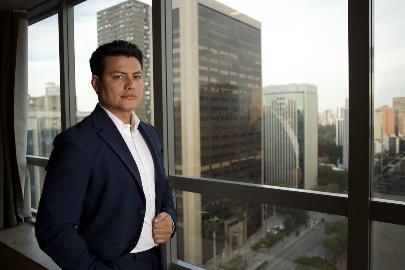 Uemerson Gabriel de terno, diante da janela de um escritório com prédios ao fundo